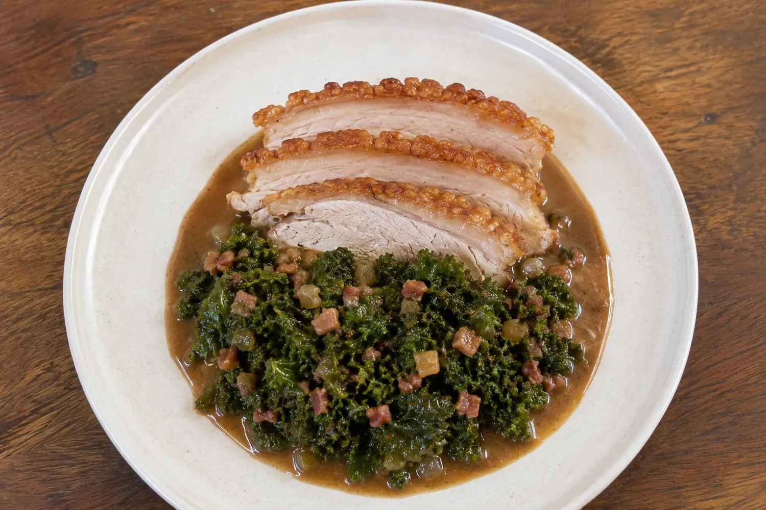 Tre skiver dansk flæskesteg med sprød svær, serveret med grønkål, små baconstykker, sauterede løg og brun keto-sauce.