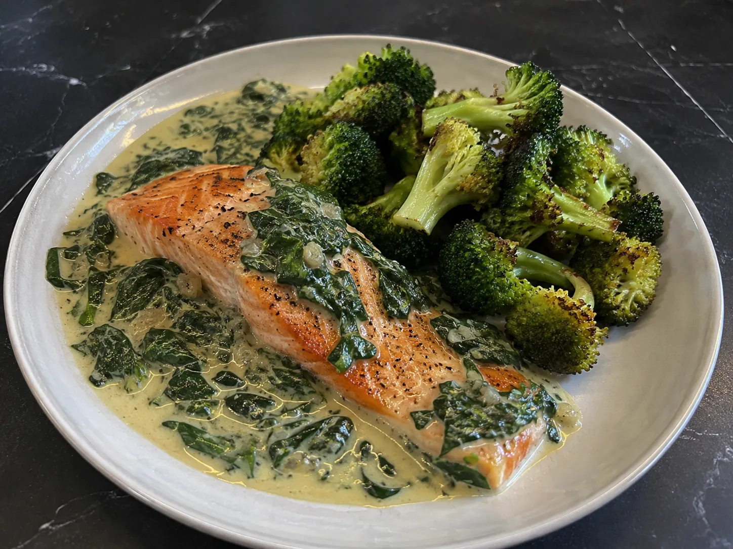 Tallerken med saftig keto laks i cremet spinatsauce serveret med smørstegt broccoli på et mørkt gråt marmorbord.