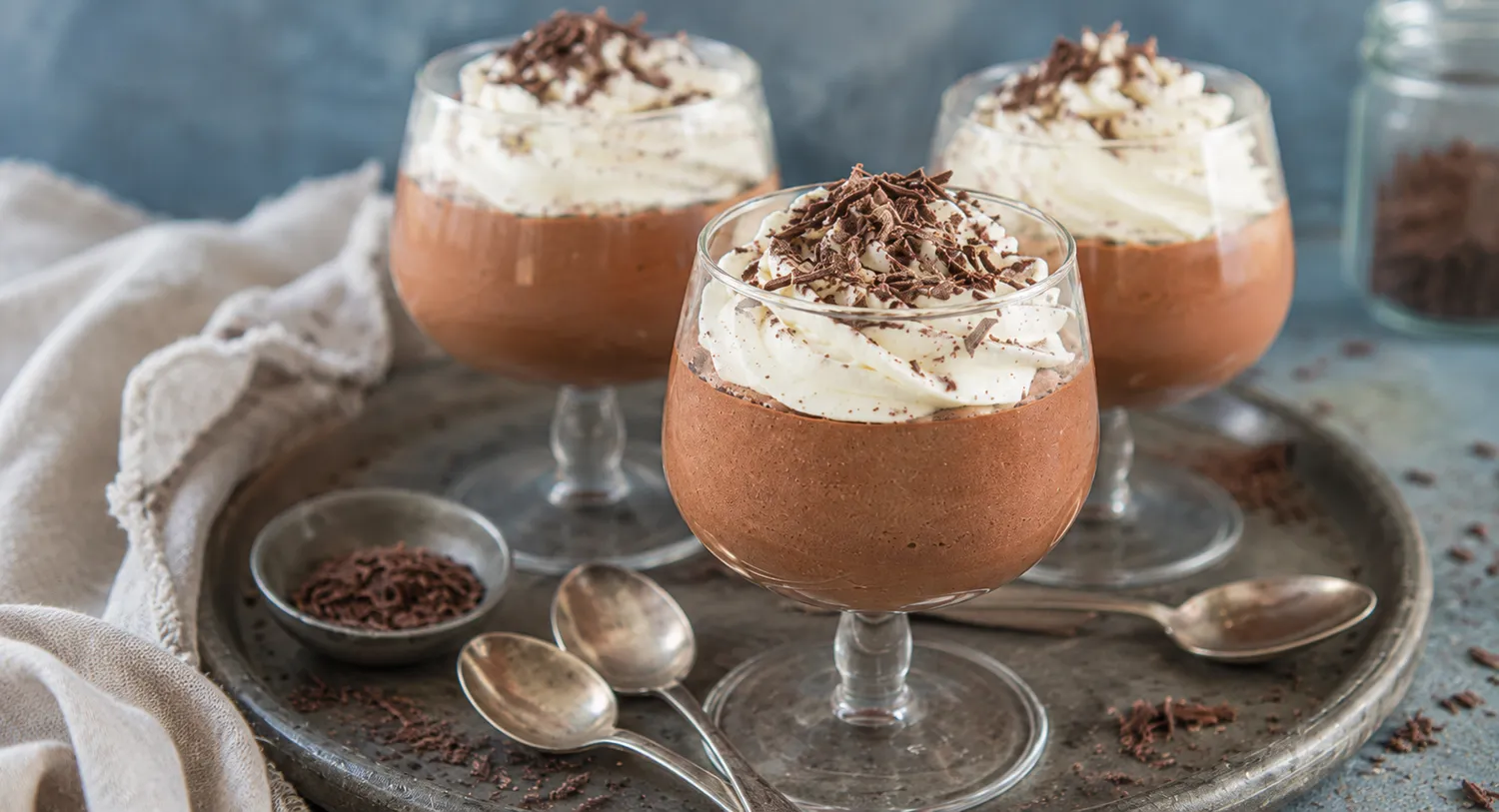 Tre glas med keto chokolademousse toppet med flødeskum og chokoladespåner på en mørk bakke med skeer