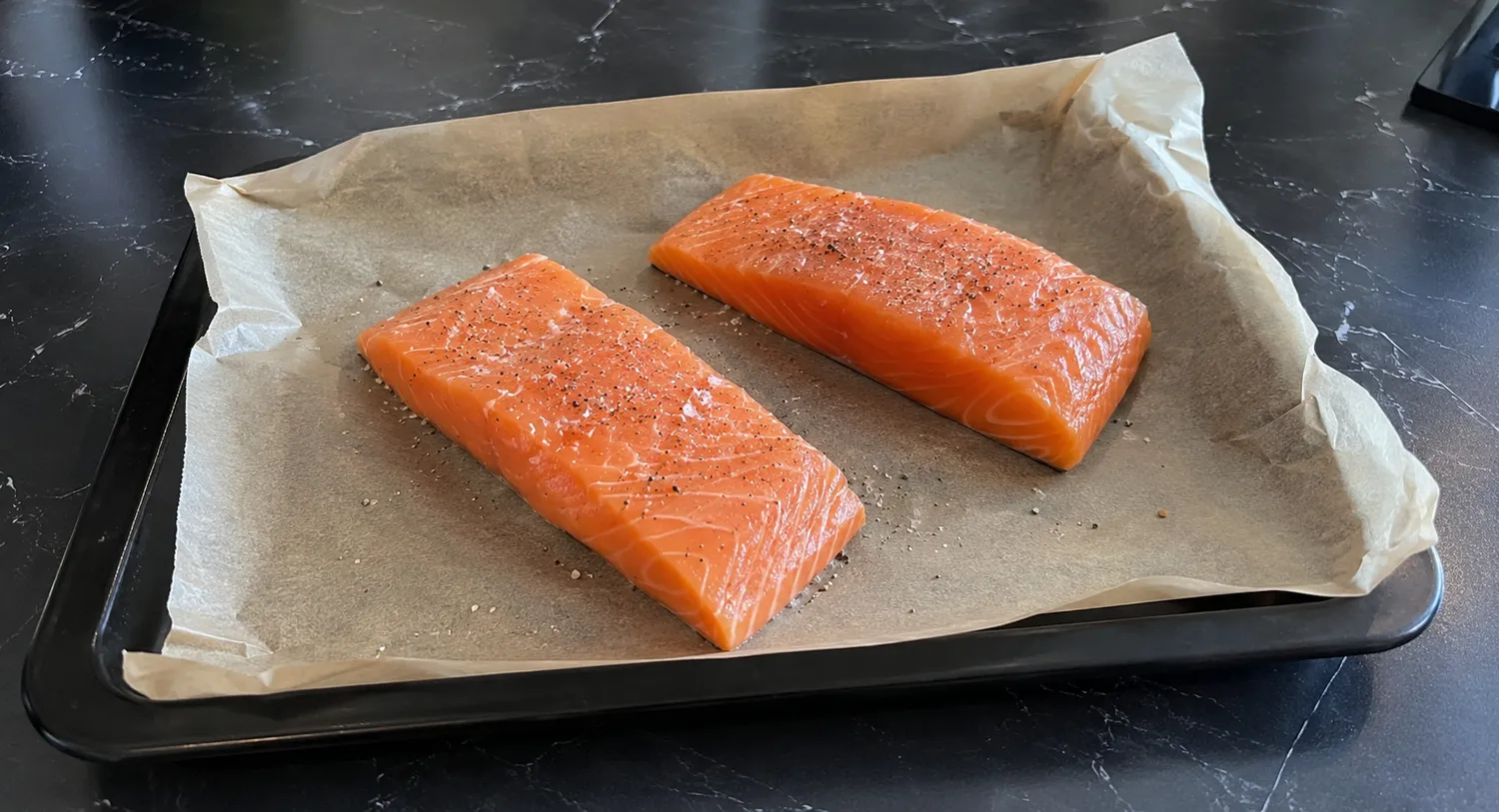 To rå laksfileter krydret med salt og peber på en bageplade med bagepapir.