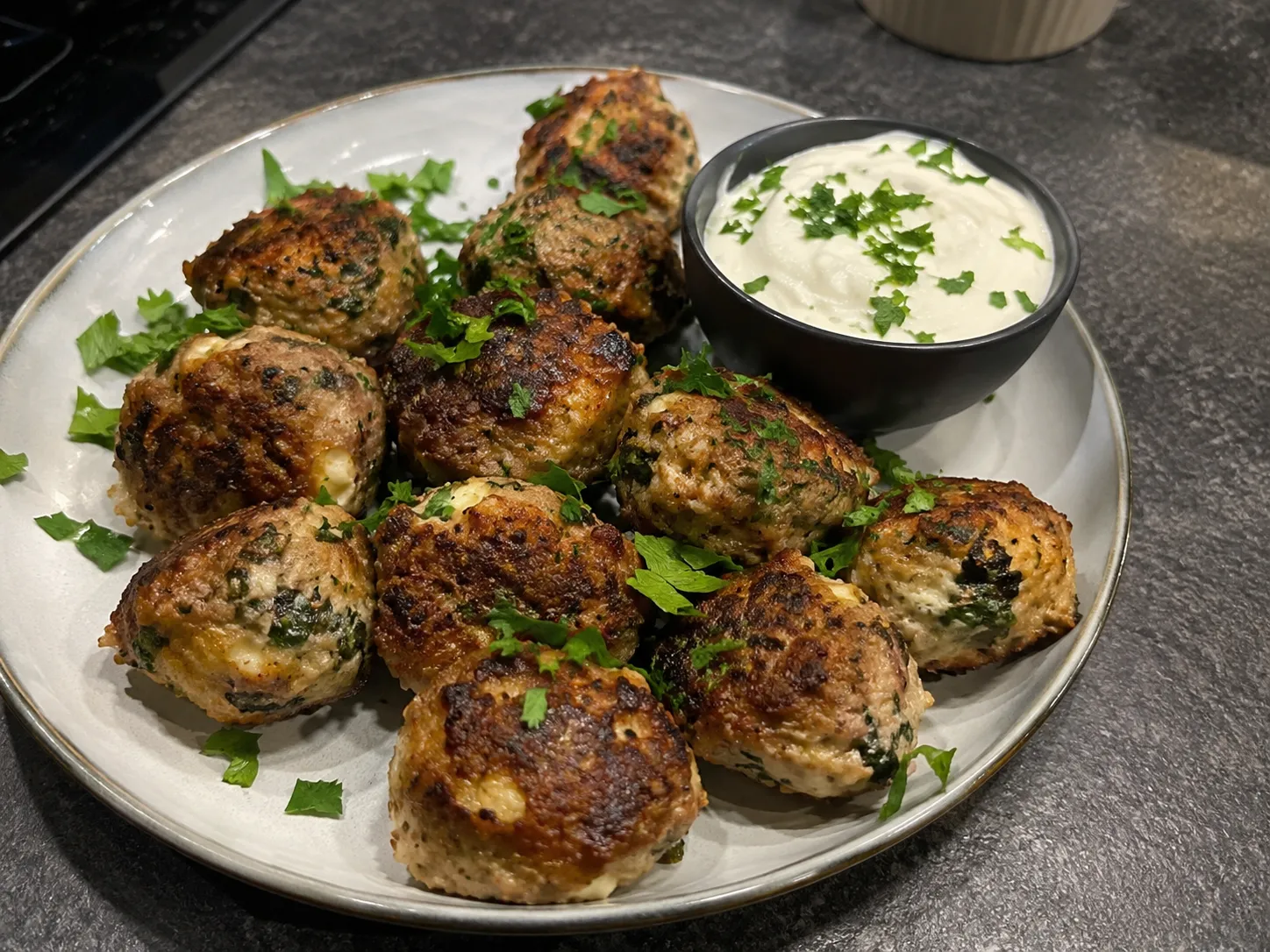 Tallerken med saftige keto kyllingekødboller med ost og spinat, pyntet med persille og serveret med en cremet dip på et mørkt gråt marmorbord.